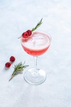 Vibrant pink cocktail, adorned with fresh raspberries and a sprig of rosemary, sits in a stylish coupe glass, ready to be enjoyed