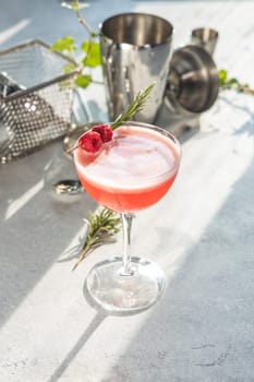 Pink cocktail decorated with raspberries and rosemary sprig, stands on bright table with cocktail shaker and jigger in background