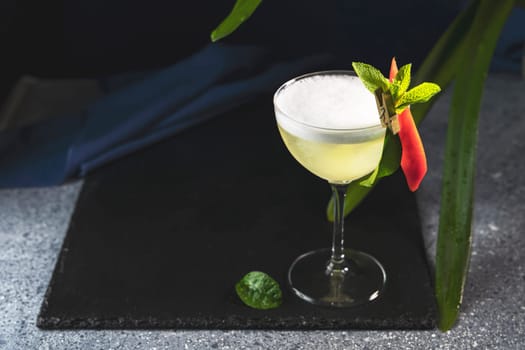 Yellow cocktail is served in a coupe glass, garnished with red pepper, mint, and egg white foam, placed on a black slate