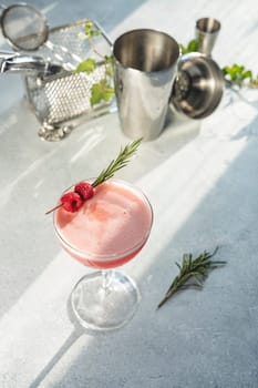 Pink cocktail is garnished with fresh raspberries and rosemary sprig on light gray background with cocktail shaker and jigger out of focus