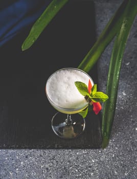 Pisco sour cocktail is being served on a black slate, decorated with fresh mint and exotic flower