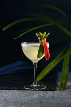 Yellow cocktail garnished with a chili pepper and mint leaves, served in a coupe glass on a dark background with tropical plant leaves