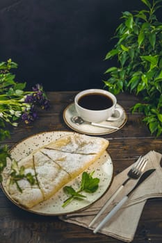 Apple strudel sprinkled with icing sugar, served on a plate next to a cup of coffee and cutlery on a wooden table