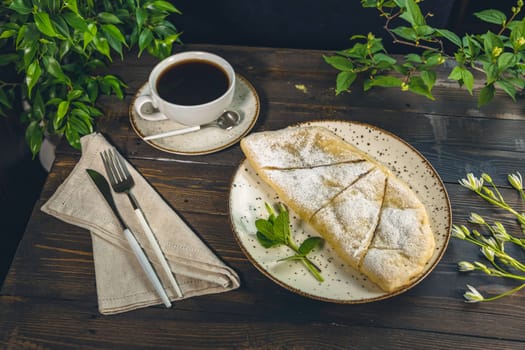 Delicious apple strudel with powdered sugar served on a plate with a cup of coffee, cutlery and napkin on a wooden table