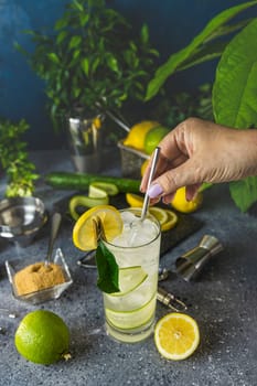 Hand adding metal straw to glass of refreshing homemade lemonade with cucumber, lime, lemon, ice and mint on grey table with ingredients and bar tools