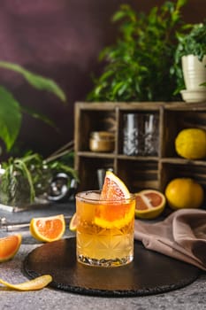 Glass of vibrant cocktail featuring grapefruit, ice, and a stylish garnish, placed on a bar counter with various ingredients and bar tools in the background