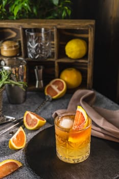 Vibrant cocktail sits on a bar counter, garnished with a slice of grapefruit and ice, with bar equipment and fresh citrus fruits in the background