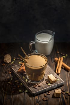 Glass of aromatic coffee with milk, cinnamon sticks, cardamom, coffee beans and brown sugar cubes on wooden table