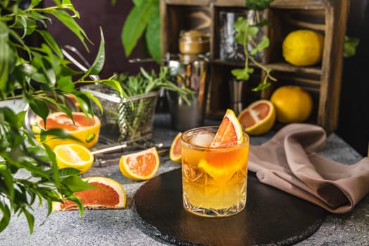 Glass of refreshing orange paloma cocktail is decorated with grapefruit slice and ice cubes, surrounded by fresh citrus fruits and green plants