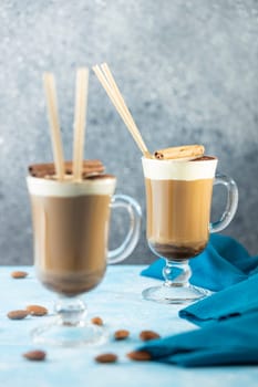 Refreshing coffee drinks topped with whipped cream, cinnamon sticks, and chocolate powder, served on a blue table with scattered almonds