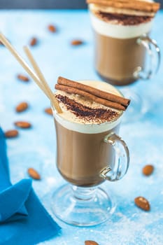Two glasses of rich coffee topped with whipped cream, cinnamon sticks, and chocolate powder, set against a light blue background