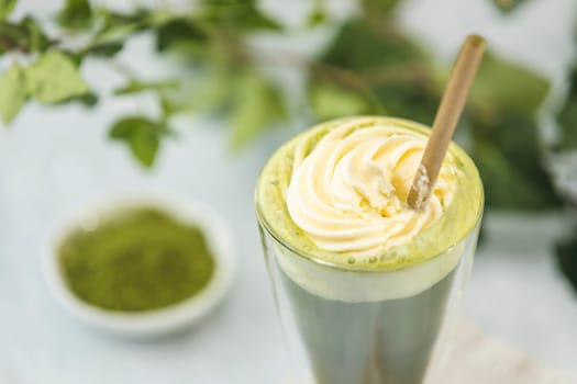 Glass of matcha latte topped with whipped cream and accompanied by powdered green tea on a blurred background with green leaves