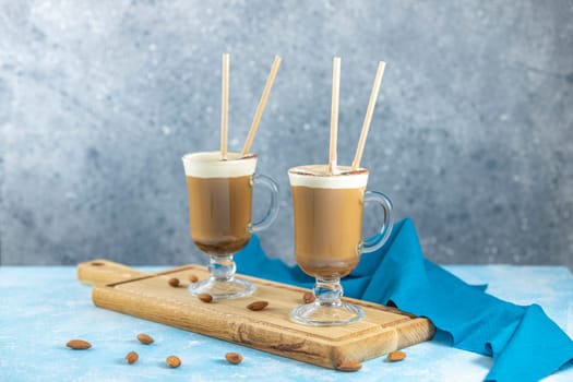 Two glasses of creamy almond coffee, complete with straws, sit on a wooden board, accompanied by scattered almonds and a blue napkin