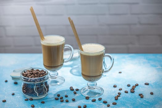 Two glasses of latte with wooden stirrers and coffee beans on a blue table create a cozy and inviting coffee break scene
