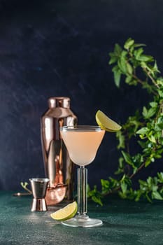 Refreshing pink cocktail is being poured into elegant glass, garnished with lime wedge, with copper shaker and jigger on dark background
