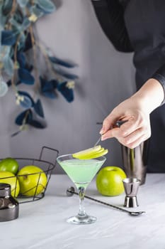 Bartender adding apple slices to a green cocktail in a martini glass, with bar equipment and fresh apples nearby