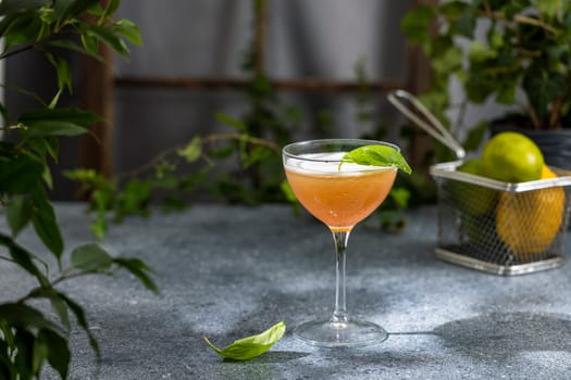 Vibrant pink cocktail garnished with fresh basil leaf resting in a stemmed glass on a gray surface, surrounded by citrus and greenery