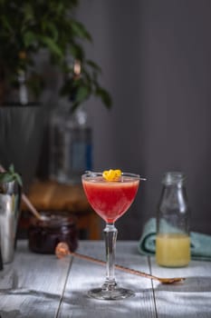 Vibrant red cocktail sits in a stemmed glass, garnished with a lemon twist, on a bar counter with cocktail making tools and ingredients