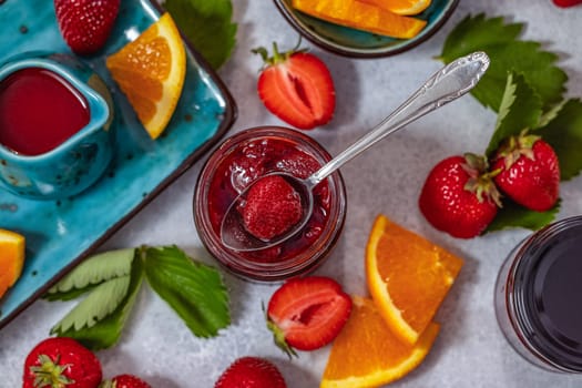 Spoon picking a strawberry from a jar of homemade strawberry jam surrounded by fresh strawberries, orange slices, strawberry sauce, and green leaves