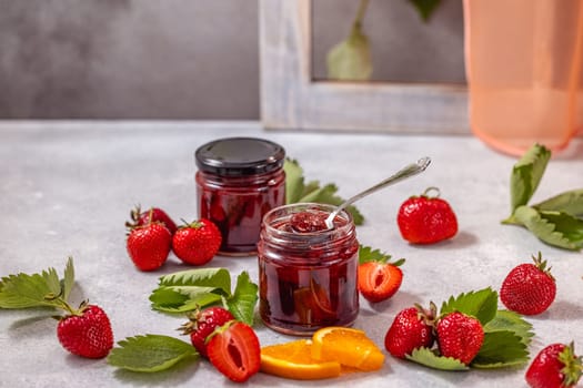 Two jars of strawberry jam surrounded by fresh strawberries, orange slices, and leaves, creating a vibrant and appetizing scene