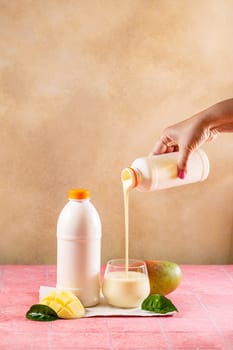 Hand pouring mango lassi or yogurt drink from bottle into glass with fresh mango fruit and leaves on pink tiled table