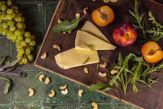 Gourmet cheese selection with fresh grapes, plums, apricots, cashew nuts, and aromatic herbs, creating an enticing culinary scene on a rustic wooden board