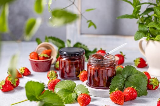 Two jars of strawberry jam, one with a spoon inside, surrounded by fresh strawberries, leaves, and a mint plant