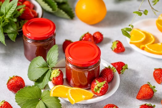 Two glass jars with red lids filled with strawberry jam, accompanied by fresh strawberries and orange slices on a white table