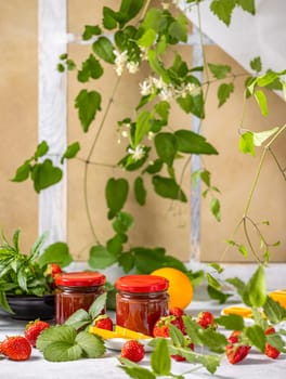 Homemade strawberry jam in glass jars with fresh strawberries, orange slices, mint leaves, and climbing plant on a table