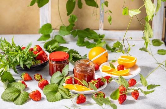 Two jars of homemade strawberry jam surrounded by fresh strawberries, orange slices, mint leaves and a climbing plant on a bright surface