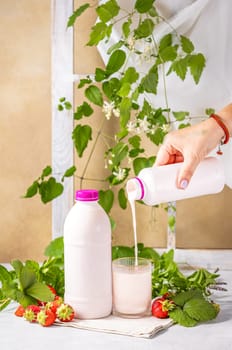 Hand pouring strawberry yogurt from bottle into glass, with fresh strawberries, mint, and decorative plants, promoting healthy eating and refreshment