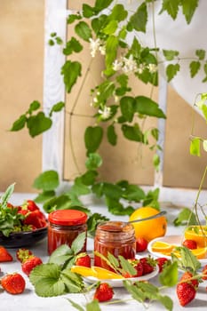 Jars of strawberry jam surrounded by fresh strawberries, orange slices, and green leaves create a vibrant and appetizing scene