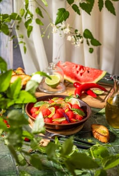Watermelon salad with lime, herbs, and chili pepper is served on a table with toasted bread, olive oil, and fresh ingredients, creating a vibrant summer scene