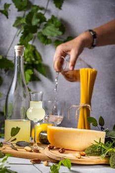Chef pouring white wine into glass, next to aged cheese, spaghetti, limoncello, brazil nuts, rosemary, and sage on wooden cutting board