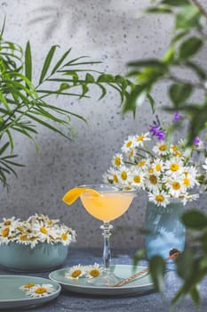Vibrant yellow cocktail, garnished with an orange twist, sits on a table adorned with daisies and lush tropical plants, creating a refreshing summer scene
