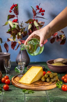 Woman pours vodka from a green bottle into a small glass on a table set with cheese, pickles, tomatoes, and other snacks