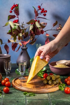 Hand placing cheese on a wooden board with traditional russian snacks like pickles, cherry tomatoes, and vodka shots