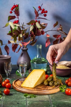 Hand placing a gherkin on a wooden board with cheese, accompanied by vodka shots, cherry tomatoes, and couscous, creating a rustic still life