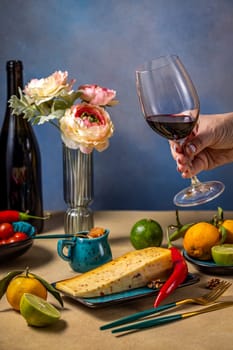 Hand holding a glass of red wine over a table set with cheese, fruits, vegetables, and flowers, creating a sophisticated and inviting atmosphere