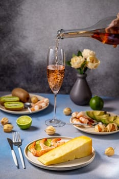 Pouring rose wine into a glass alongside a vibrant assortment of cheese, shrimp, kiwi, lime, avocado, and physalis