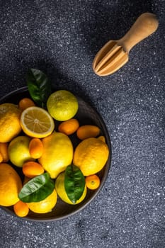 Vibrant citrus fruits like lemons, limes, and kumquats arranged in a black bowl alongside a wooden reamer, creating a refreshing scene on a dark stone tabletop