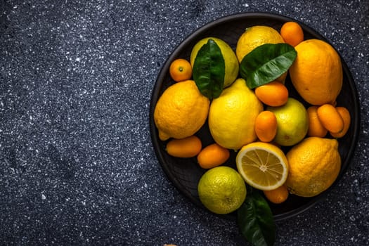 Vibrant citrus fruits, including lemons, limes, kumquats, and oranges, create a colorful arrangement on a dark plate, showcasing their freshness and variety