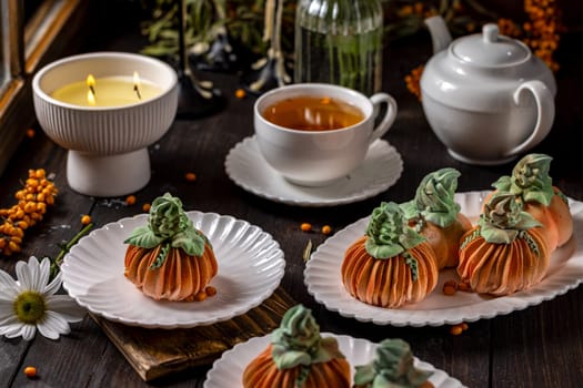 Pumpkin shaped desserts are served on white plates alongside a cup of tea and a burning candle, evoking a warm and inviting autumnal ambiance