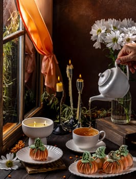 Cozy autumn scene with pumpkin shaped desserts, a woman pouring tea, and candles by a window on a rainy day