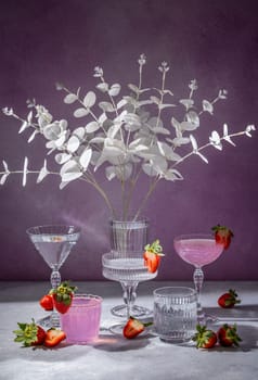 Pink cocktails and elegant glassware are beautifully arranged with fresh strawberries and white eucalyptus branches, creating a stylish and refreshing scene