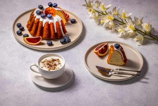 Gugelhupf cake with white icing, blueberries and blood orange slice, served with cappuccino and orchid decoration, creates a tempting table setting