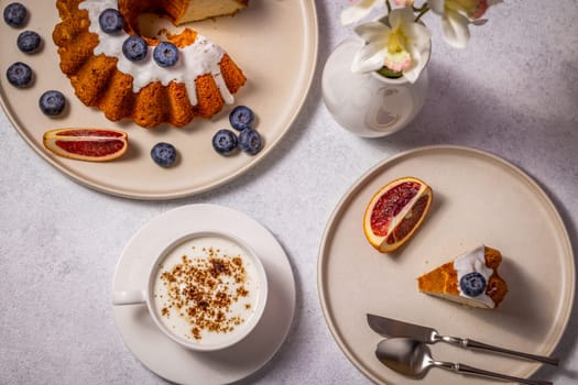 Flat lay of a delicious homemade cake with icing and blueberries, accompanied by a cappuccino and blood orange slices, perfect for a delightful breakfast