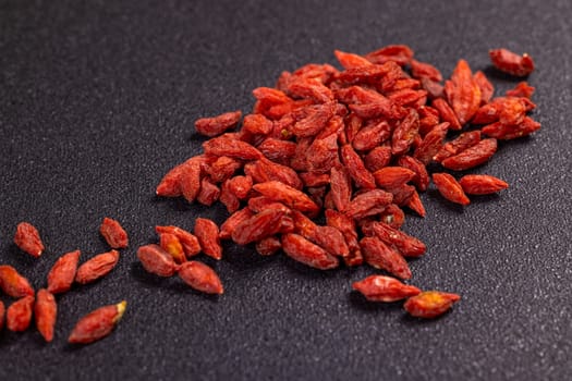 Numerous dried goji berries are scattered across a dark surface, creating a visually appealing arrangement