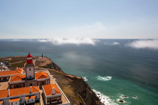 Drone view of Cabo da Roca lighthouse perched on cliff overlooking seascape in Portugal. The red roofed lighthouse stands as striking landmark, highlighting dramatic beauty of coastal location