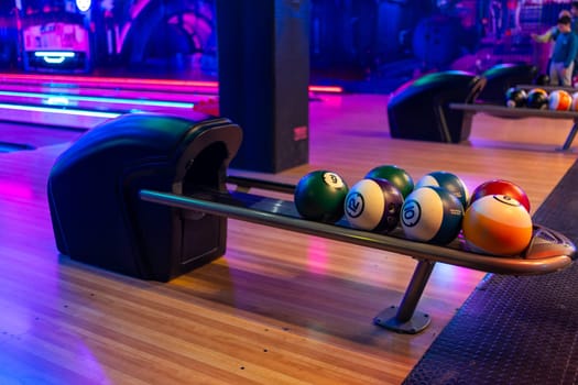 Various bowling balls arranged in stand on wooden lane at bowling hall. Illuminated playroom in shopping center with no people